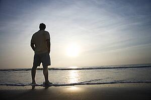 Caucasian mid-adult man standing alone on beach looking at ocean at sunrise.