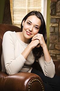 Caucasian/Hispanic young woman sitting in leather chair looking at viewer smiling with head on hands.