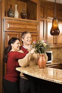 Caucasian woman leaning on Caucasian man while he arranges flowers in vase in kitchen.