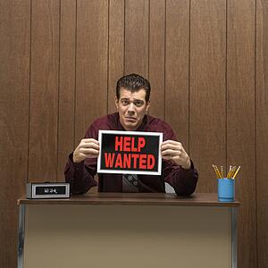 Caucasion mid-adult retro businessman with desperate expression sitting at desk holding up help wanted sign.