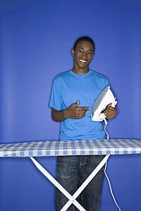 Portrait of smiling African-American teen boy pointing at iron standing at iron board against blue background.