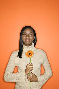 Portrait of grinning African-American teen girl holding single Gerbera Daisy against orange background.