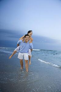 Caucasian mid-adult male carrying female piggyback style on beach.
