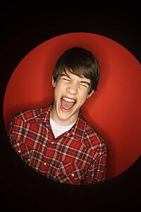Portrait of Caucasian teen boy screaming with eyes closed against red background.