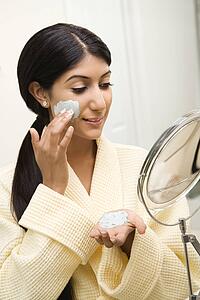 Smiling Asian/Indian young woman looking in mirror and applying facial scrub.