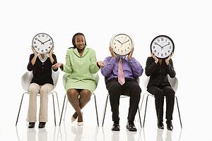 Business people sitting holding clocks over faces while African-American businesswoman shrugs.