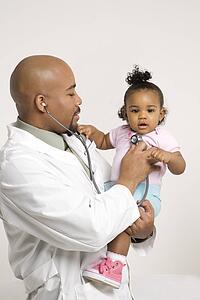 African-American male pediatrician holding and examinating baby girl with stethoscope.