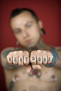 Caucasian mid-adult man with tattoos and piercings holding out fists reading beat down.