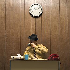 Caucasion mid-adult retro businessman wearing fedora sitting at desk drinking coffee.