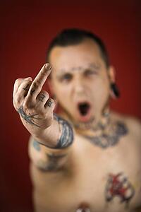 Caucasian mid-adult man with tattoos and piercings holding up middle finger.