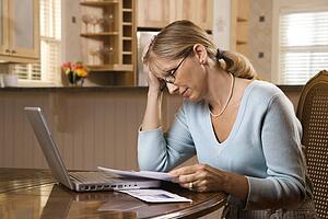 Caucasian mid-adult woman paying bills on laptop computer resting head in hand with worry.
