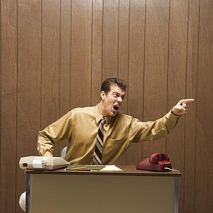 Caucasion mid-adult retro businessman sitting at desk pointing in anger.