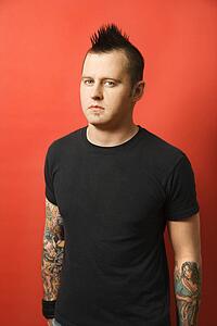 Caucasian man with mohawk and tattoos standing against orange background.