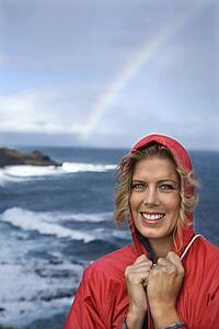 Caucasian mid-adult woman in red raincoat standing by ocean with rainbow in background in Maui, Hawaii.