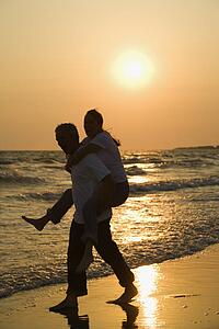 Caucasian mid-adult male carrying female piggyback on beach at sunset.