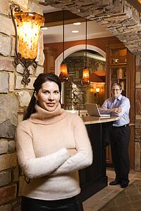 Caucasian woman standing with arms crossed looking at viewer with Caucasian man in background on laptop.