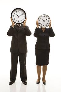 Caucasian middle-aged businessman and Filipino businesswoman holding clocks in front of their heads standing against white background.