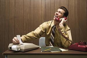 Caucasion mid-adult retro businessman sitting at desk talking on telephone using adding machine.