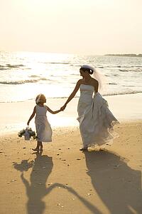 Caucasian mid-adult bride and flower girl holding hands walking barefoot on beach.