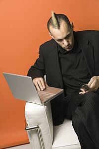 Caucasian man in suit with mohawk typing on laptop and looking at cellphone sitting in chair against orange background.