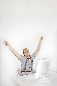 Caucasian young man dressed like nerd happy with arms raised in front of computer.