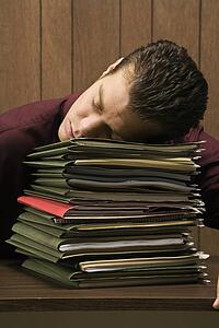Caucasion mid-adult retro businessman sitting at desk with head down sleeping on a tall stack of folders.