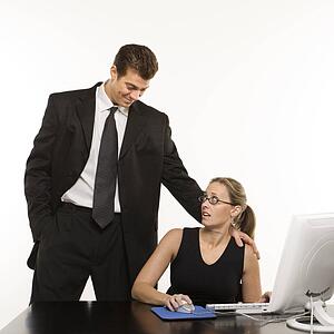 Caucasian mid-adult man touching shoulder of woman sitting at computer who feels uncomfortable.