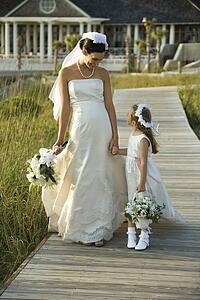 Caucasian mid-adult bride holding hands with flower girl walking down wooden beach walkway.