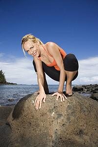 Caucasian young adult woman squatting on large rock.