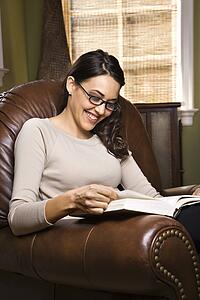 Caucasian/Hispanic young woman sitting in leather chair smiling and reading a book.