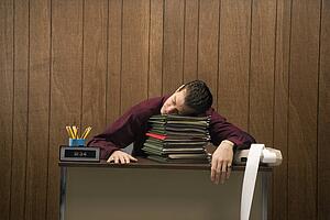 Caucasion mid-adult retro businessman sitting at desk resting his head on a tall stack of folders.