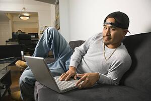 Asian young adult man wearing backwards baseball cap lounging on sofa working on laptop and looking at viewer.