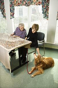 Elderly Caucasian woman and daughter putting puzzle together in retirement community center. with pet dog at their side.