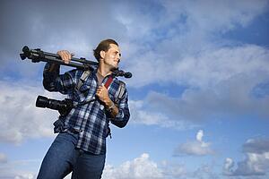 Caucasian mid-adult man standing with camera and tripod over his shoulder. 