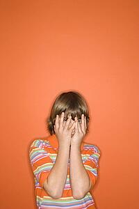 Portrait of Caucasian boy with hands covering his face standing against orange background.