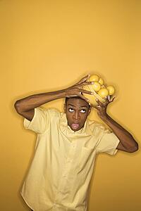 Young African-American man dropping a bowl of lemons balancing on his head, on yellow background.