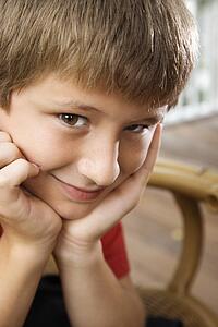 Portrait of Caucasian pre-teen boy with chin in hands looking at viewer and smiling.
