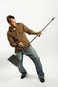 Caucasian man with mohawk playing broom like guitar against white background.