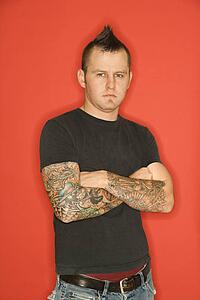 Caucasian man with mohawk and tattoos standing with arms crossed against orange background.