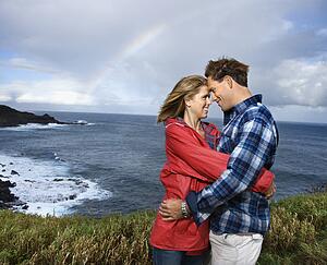 Caucasian mid-adult couple embracing by ocean with rainbow in background in Maui, Hawaii.