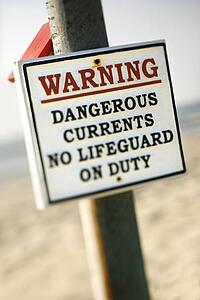 Warning sign on post at the beach with ocean in background.
