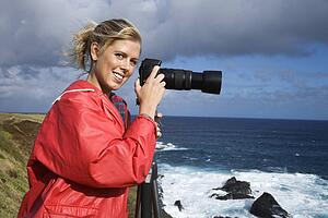 Caucasian mid-adult woman holding camera on tripod, looking at viewer, on cliff overlooking ocean in Maui, Hawaii.