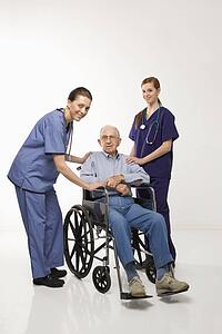 Two Caucasian females wearing scrubs with elderly Caucasian male in wheelchair.
