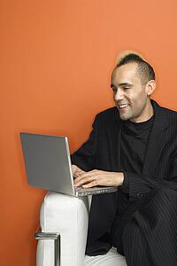 Caucasian man in suit with mohawk smiling typing on laptop against orange background.