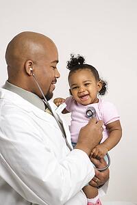 African-American male pediatrician holding and examinating baby girl with stethoscope.
