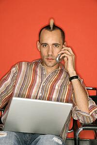 Caucasian man with mohawk holding laptop talking on cellphone against red background.