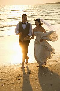 Caucasian mid-adult bride and mid-adult groom holding hands running barefoot on beach.