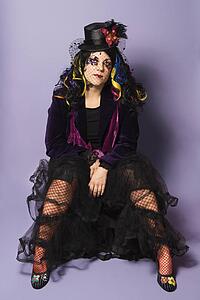 Portrait of Caucasian woman with unique hat, hair and attire sitting against purple background.