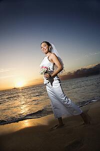 Young adult female Caucasian bride walking holding bouquet on beach.