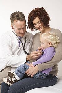Middle-aged adult Caucasian male doctor holding stethoscope to female toddler's chest with mother watching.